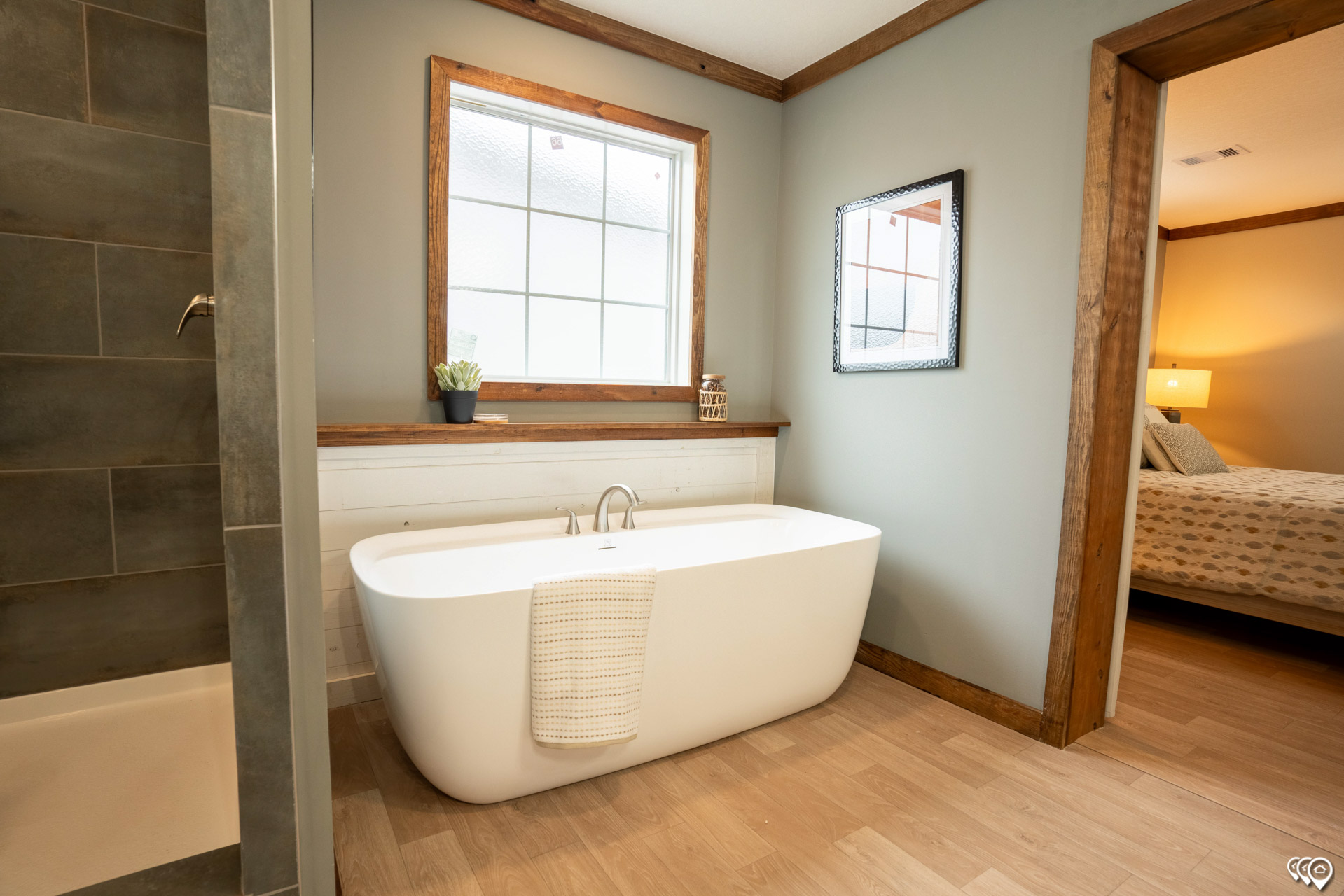 Modern bathroom with a freestanding white tub, wooden-framed window and mirror, and light wood flooring. A plush towel hangs on the tub. Calm ambiance.