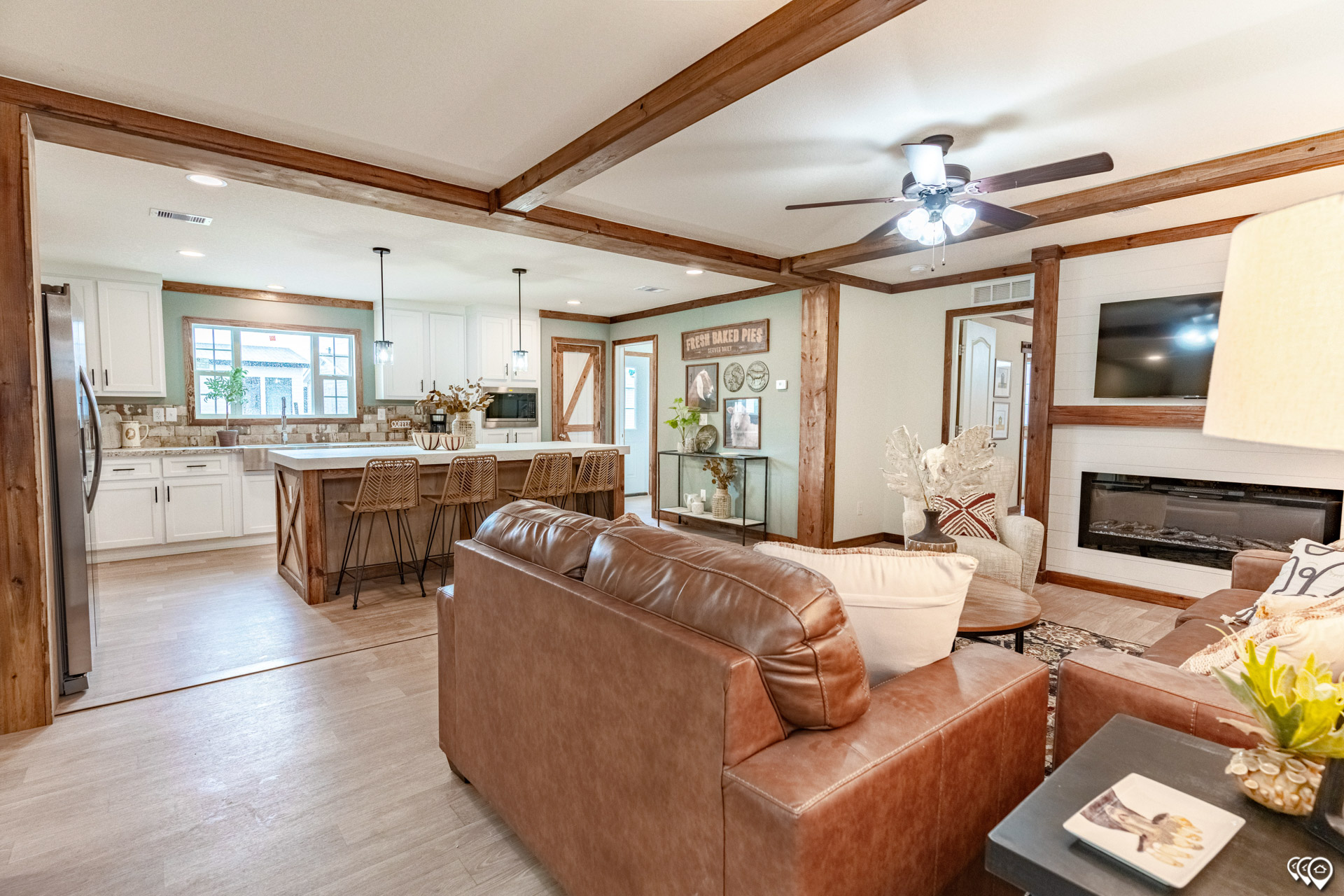 Warm, rustic open living space with wood beams and brown leather sofas. The kitchen features white cabinets, an island, and pendant lights. Cozy and inviting atmosphere.