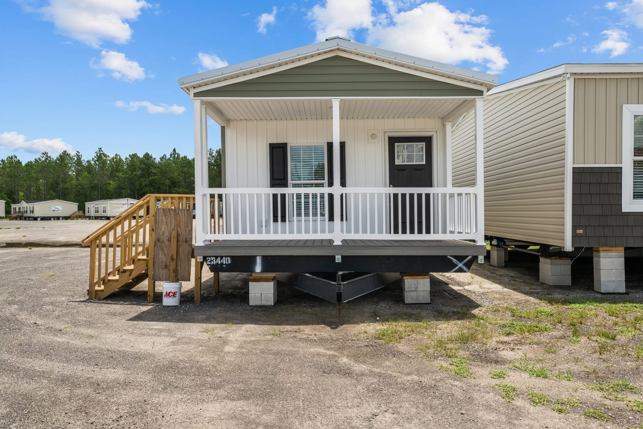 Small portable home on a gravel lot under a blue sky with clouds. It has a front porch, wooden steps, and is surrounded by other similar homes.