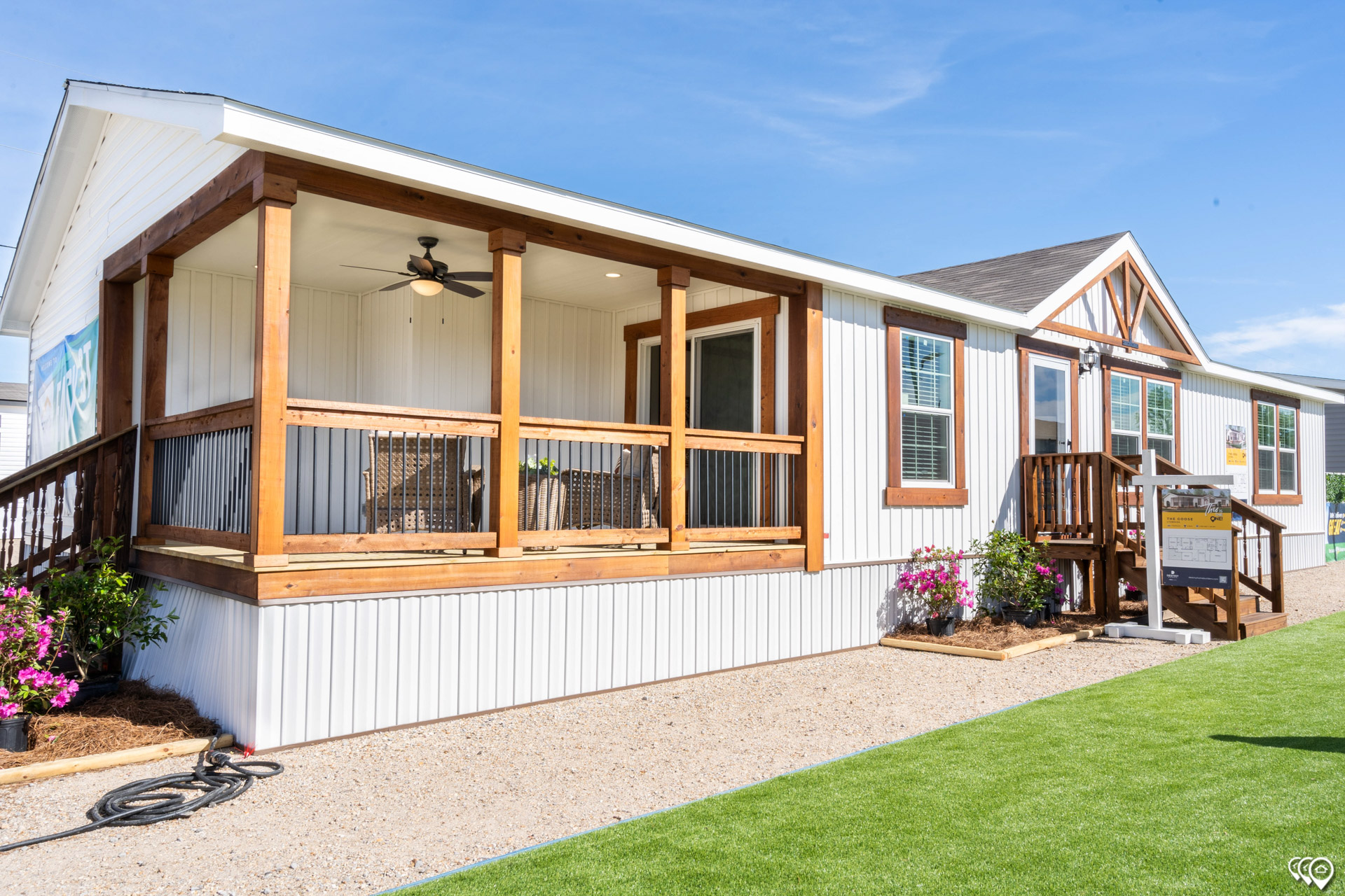 Single-story modular home with a wooden porch under a clear blue sky. The porch features a ceiling fan and chairs, surrounded by flowers and green lawn.