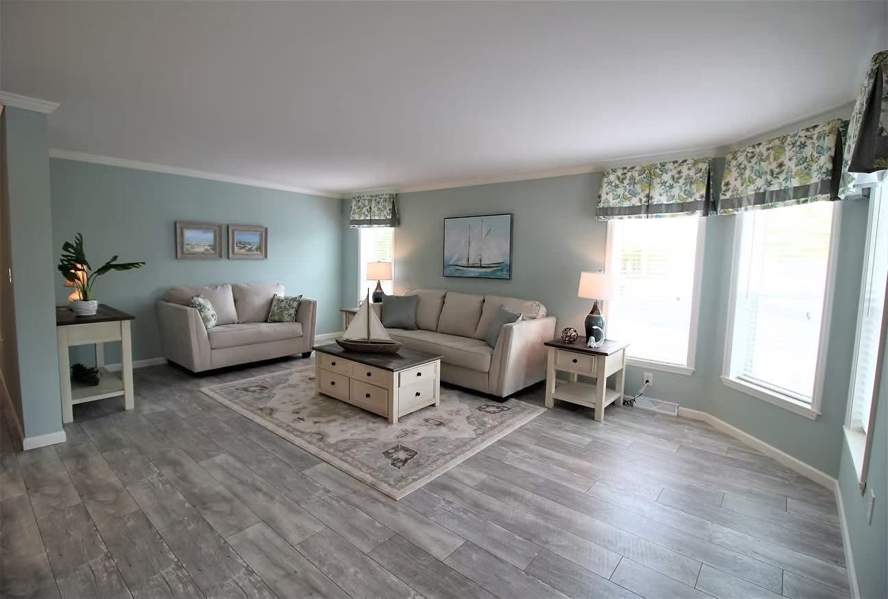 A cozy living room with light blue walls, two beige sofas, a nautical painting, and large windows with floral valances. A sailboat decorates the coffee table.