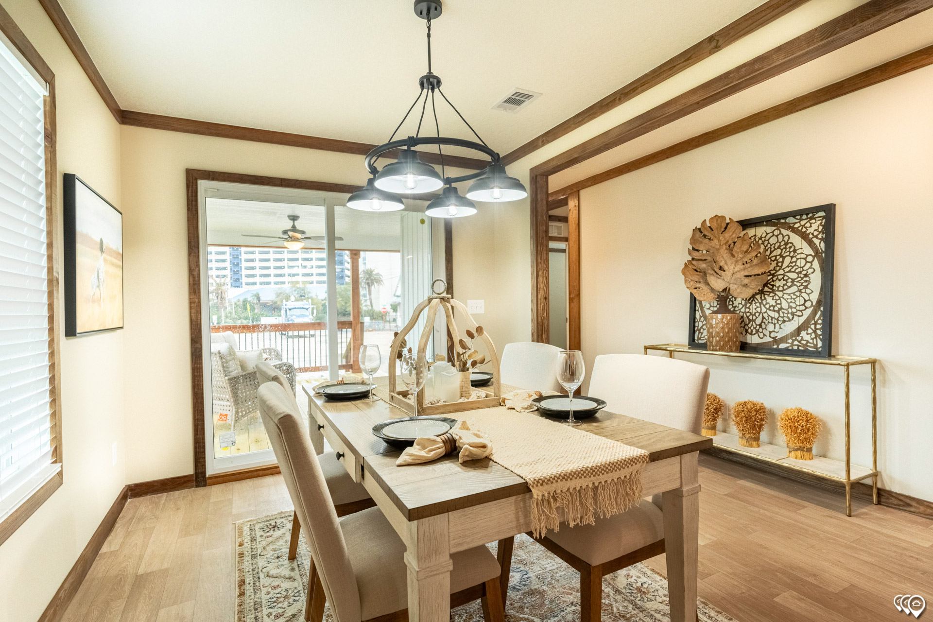 Bright dining room with a rustic wooden table set for four, featuring black plates and beige napkins. Modern chandelier above. Large window shows a patio. Neutral tones with decorative wall art and plants create a cozy ambiance.