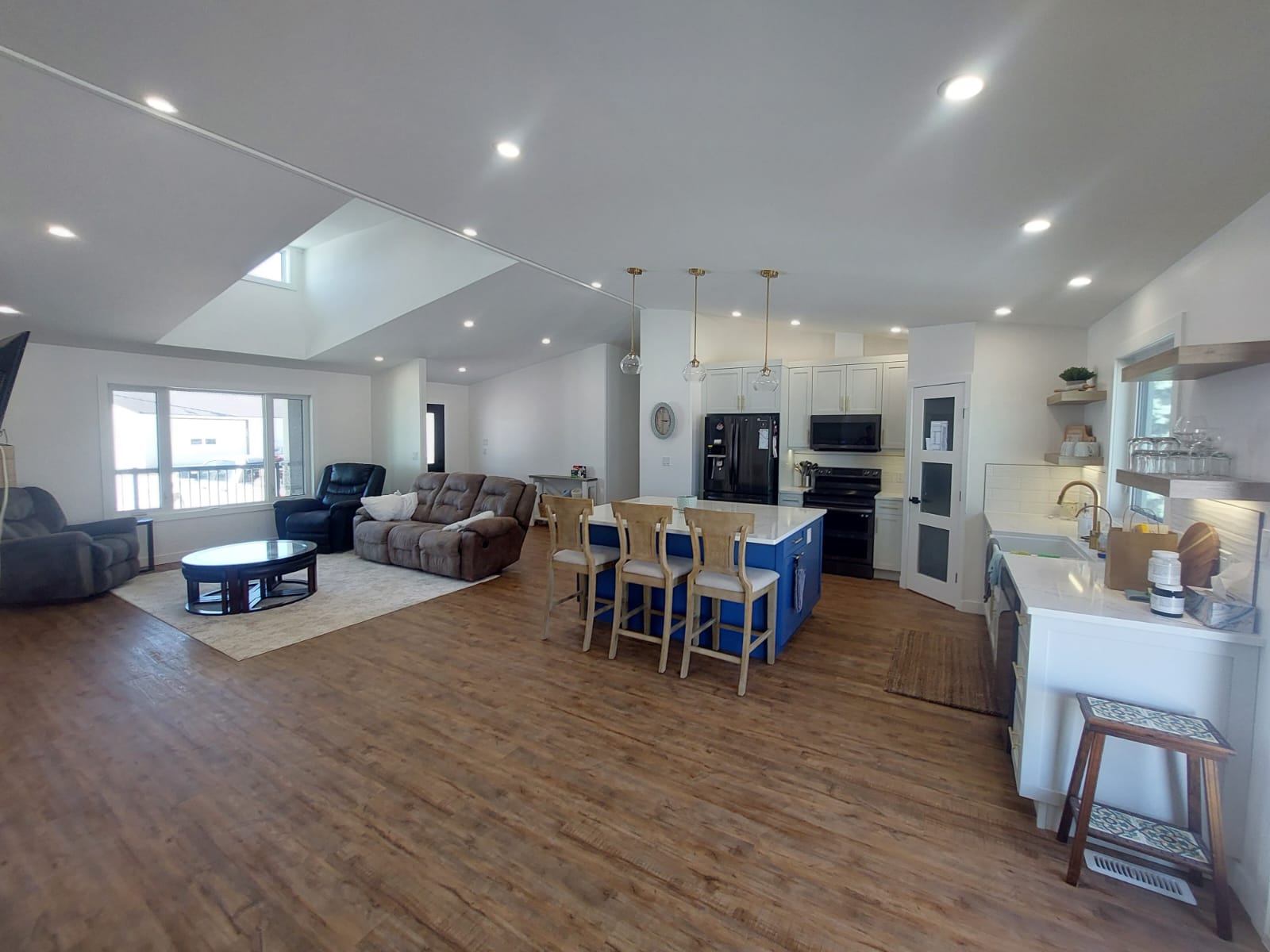 Spacious open-concept living room and kitchen with wooden floors. Features a cozy sofa, modern appliances, blue island with stools, and multiple windows.