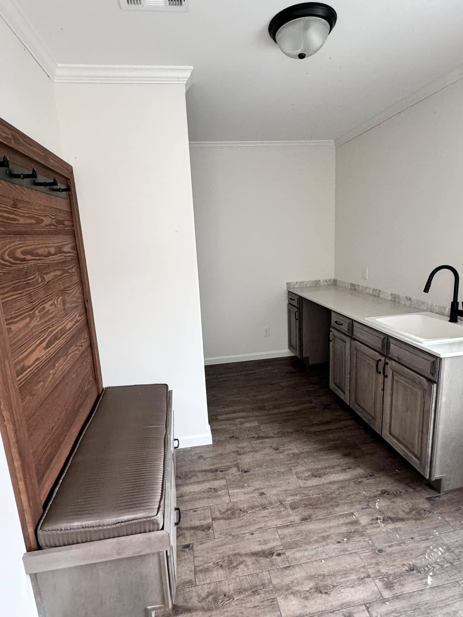 Narrow room with dark wood floor, white walls, and ceiling. Left: wooden bench with hooks. Right: small kitchen area with sink and cabinets. Minimalist and tidy setting.