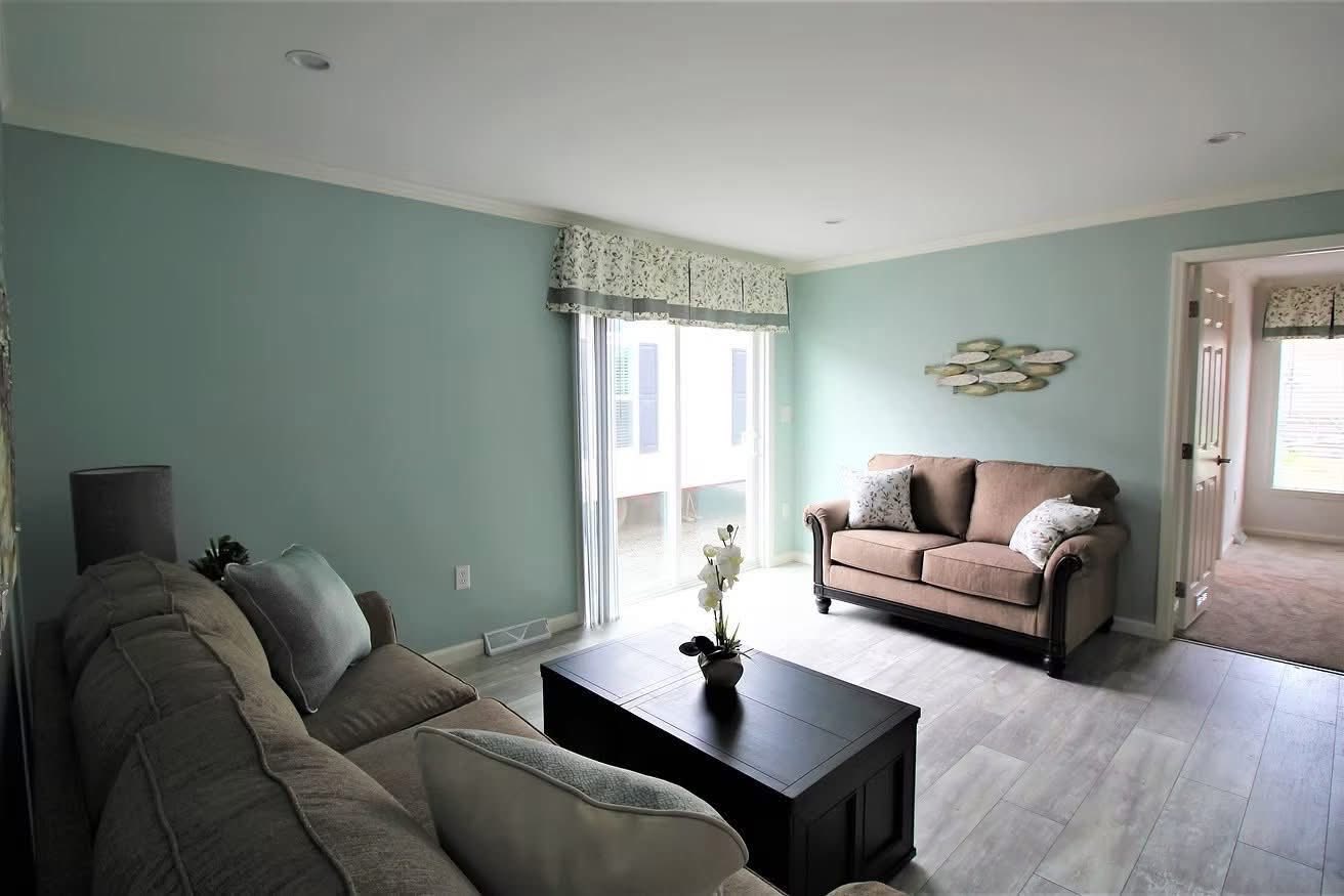 Bright living room with soft blue walls and wood flooring. Features a brown loveseat, a large dark coffee table with orchid decor, and a sofa with cushions. Light filters through sliding glass doors, creating a cozy, inviting atmosphere.