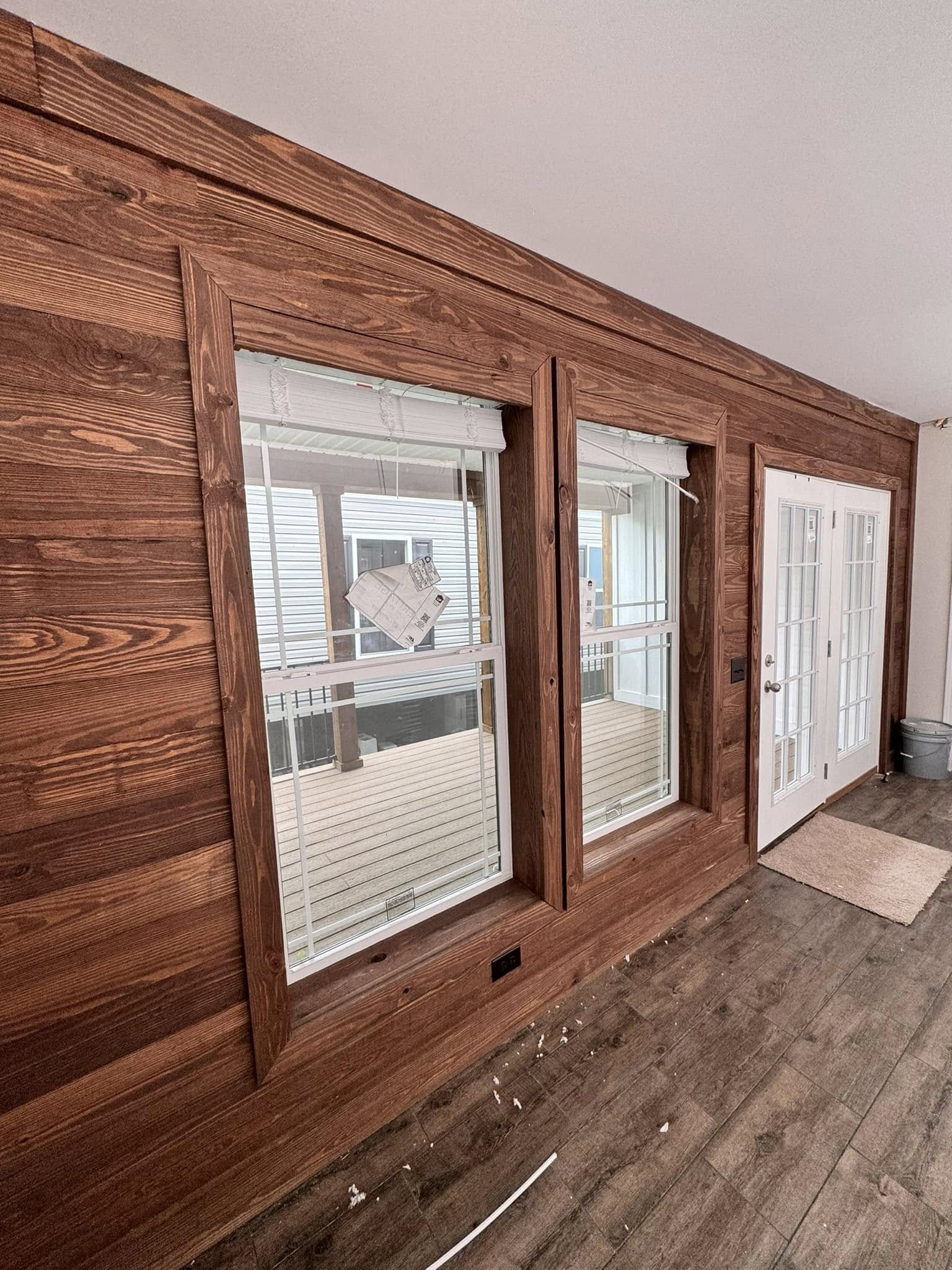 Wooden wall with two large windows and a white door, reflecting a bright and clean interior. The floor has scattered debris, suggesting recent work.