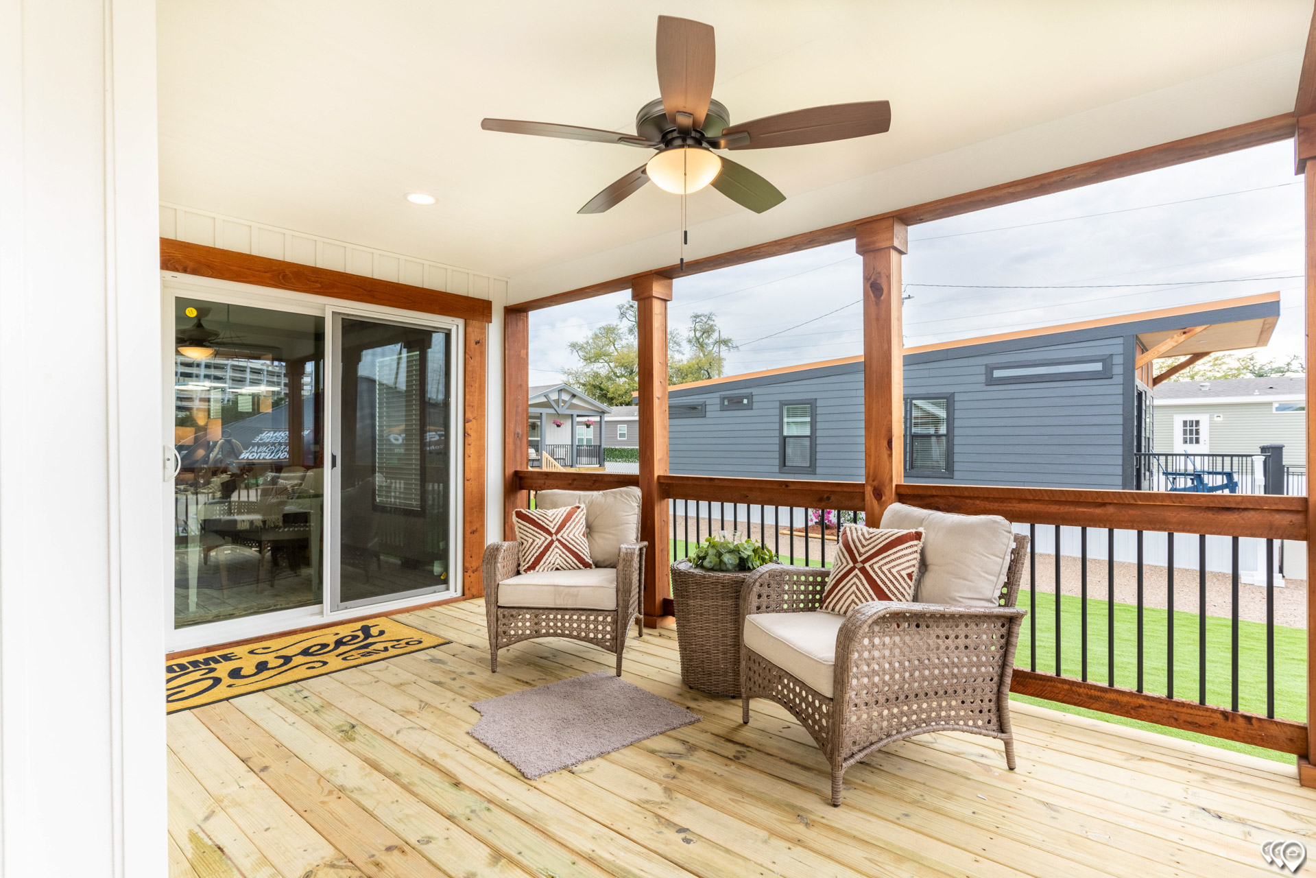 Cozy porch with wicker chairs and colorful cushions on a wooden deck. Ceiling fan above, sliding glass door, and doormat create a welcoming vibe.