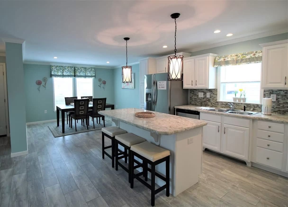 Spacious kitchen with light blue walls, white cabinets, and granite countertops. An island with three stools is lit by pendant lights. Dining area in the background.