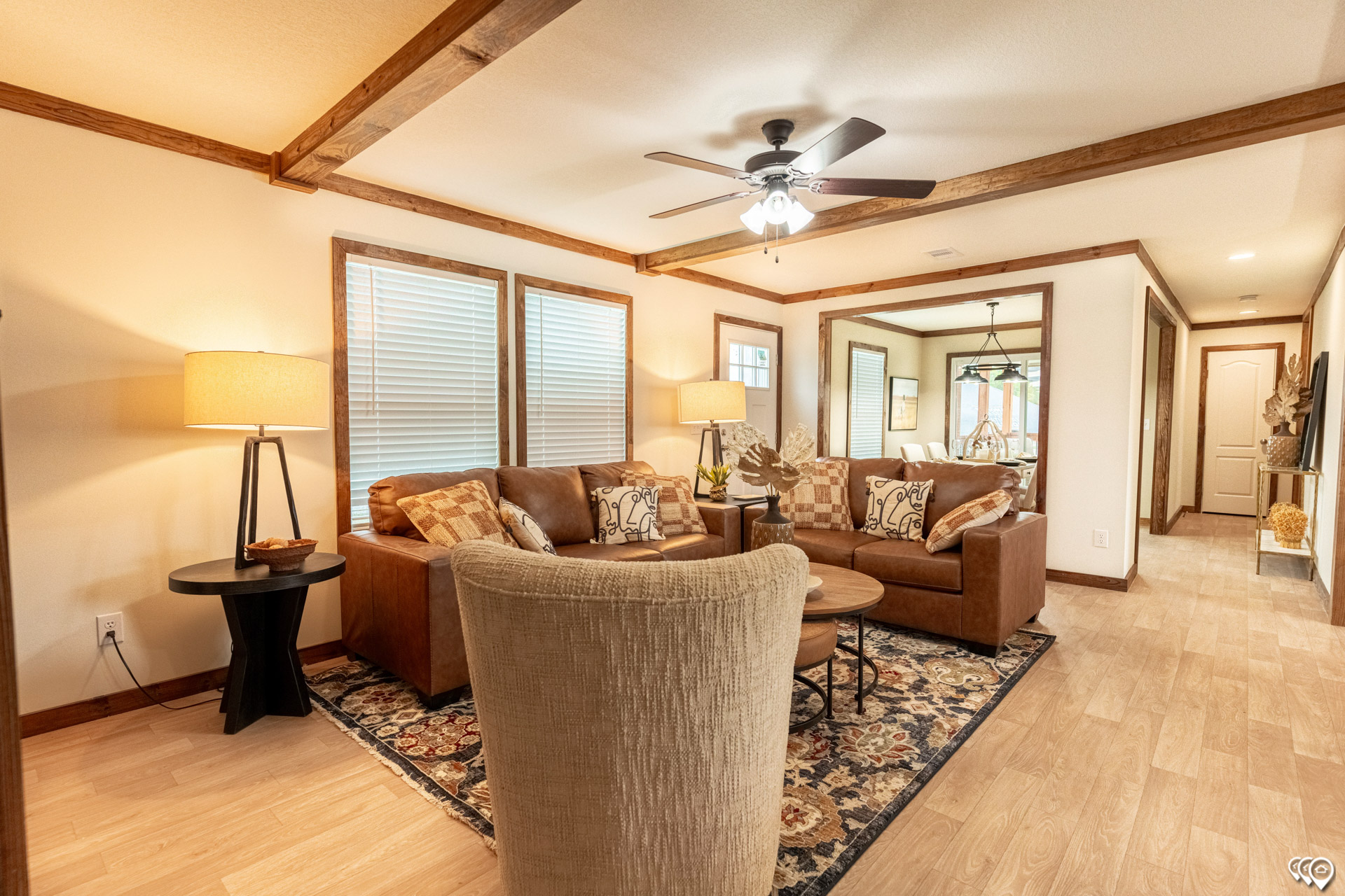 Cozy living room with leather sofas, patterned cushions, a beige armchair, and dark wood accents. Warm lighting, ceiling fan, and floral rug create a welcoming feel.