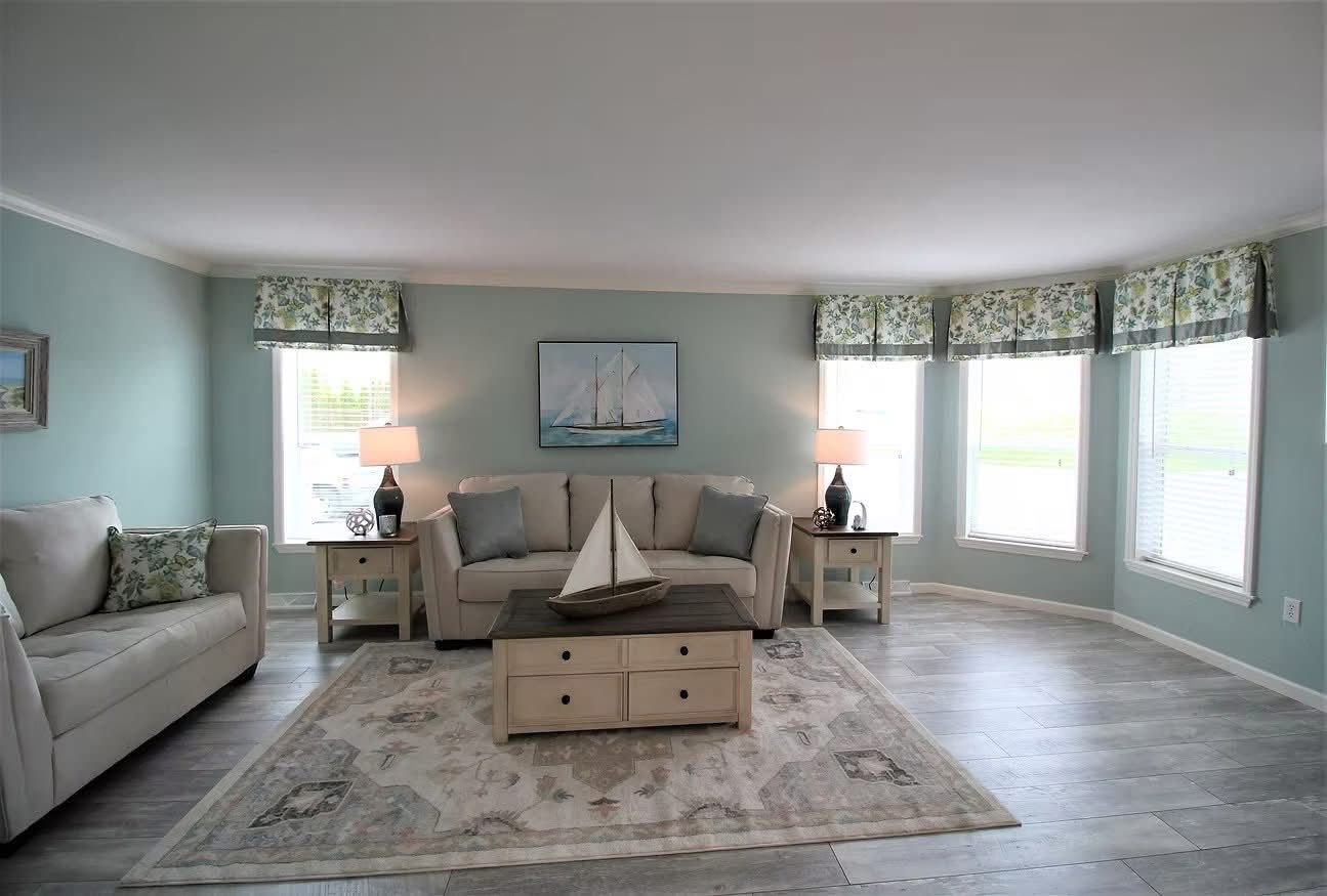 Bright living room with pale blue walls features two beige sofas, floral curtains, and nautical decor. A sailboat model sits on a rustic coffee table.