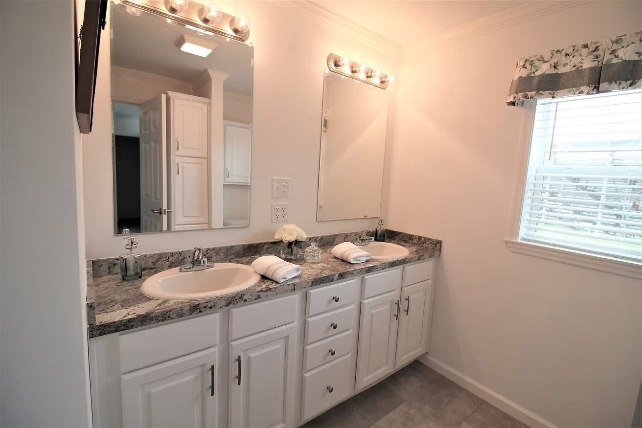 Bright bathroom featuring a dual-sink vanity with granite countertops, mirrors, and light fixtures. White cabinets and a window with floral curtains add a cozy feel.