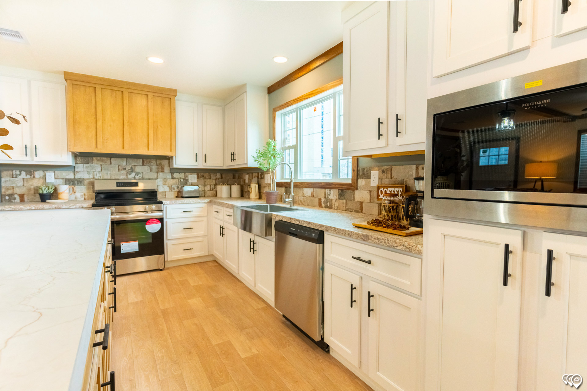 Bright kitchen with wooden flooring, white cabinets, stainless steel appliances, a brick backsplash, and a large window. Clean, modern, and inviting.