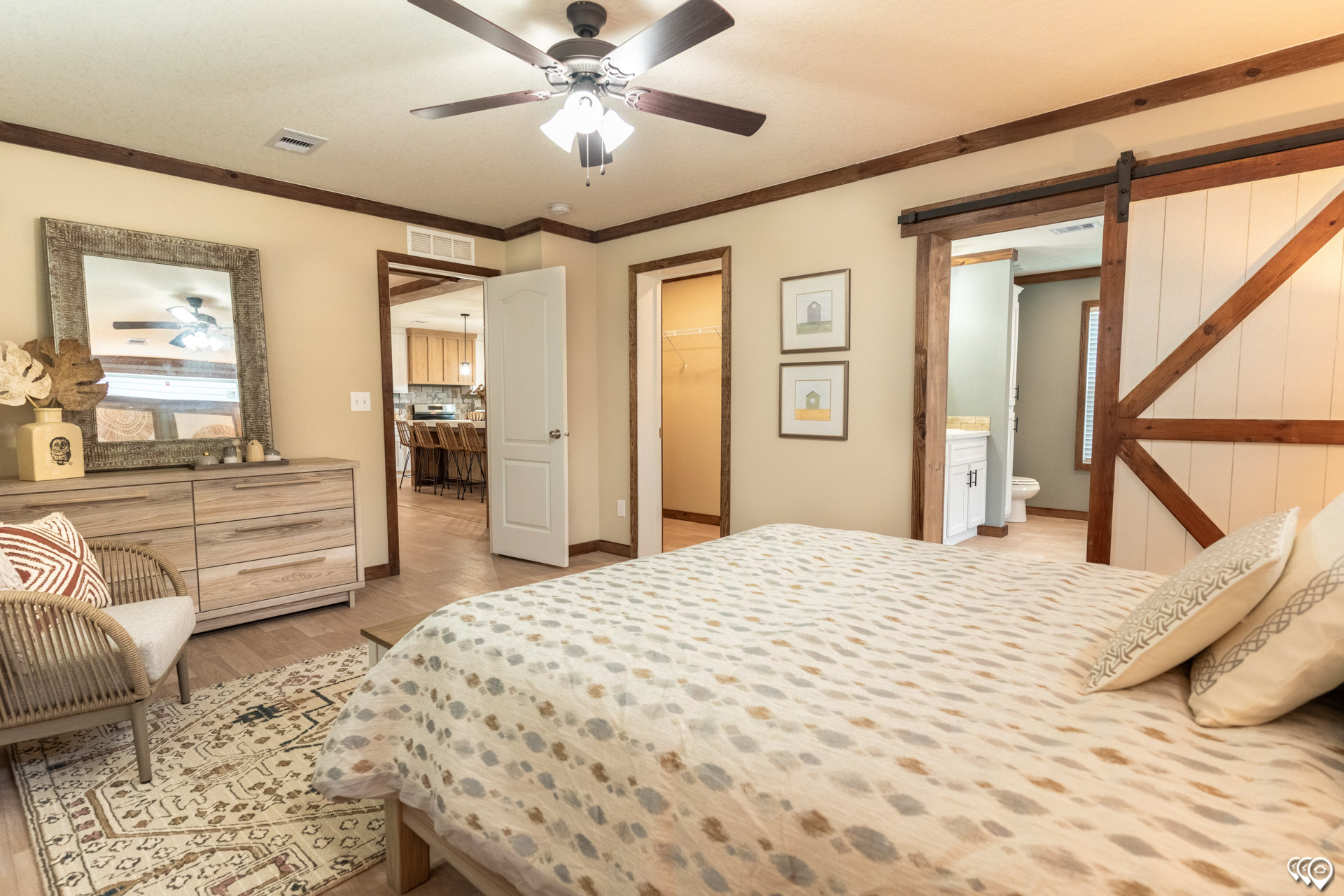 A cozy bedroom with a bed covered in a dotted blanket, wooden furniture, a barn door, ceiling fan, and neutral tones, creating a warm, inviting atmosphere.