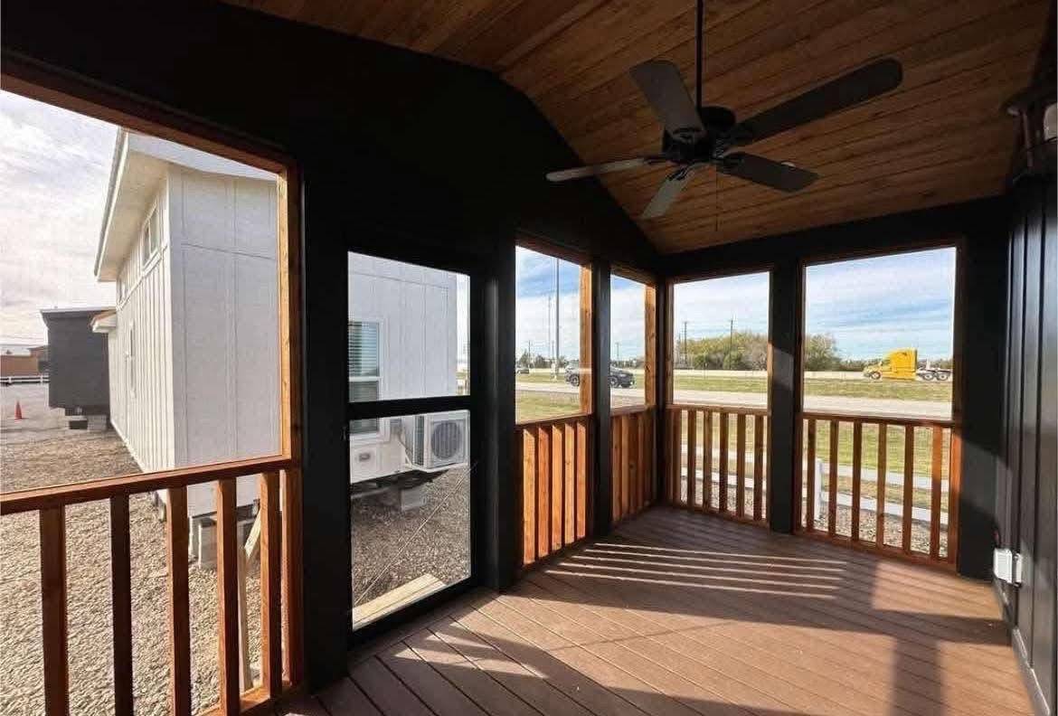 A cozy wooden porch with a ceiling fan, enclosed by railings and screens, overlooks a road and vehicle. Sunlight casts warm shadows across the floor.