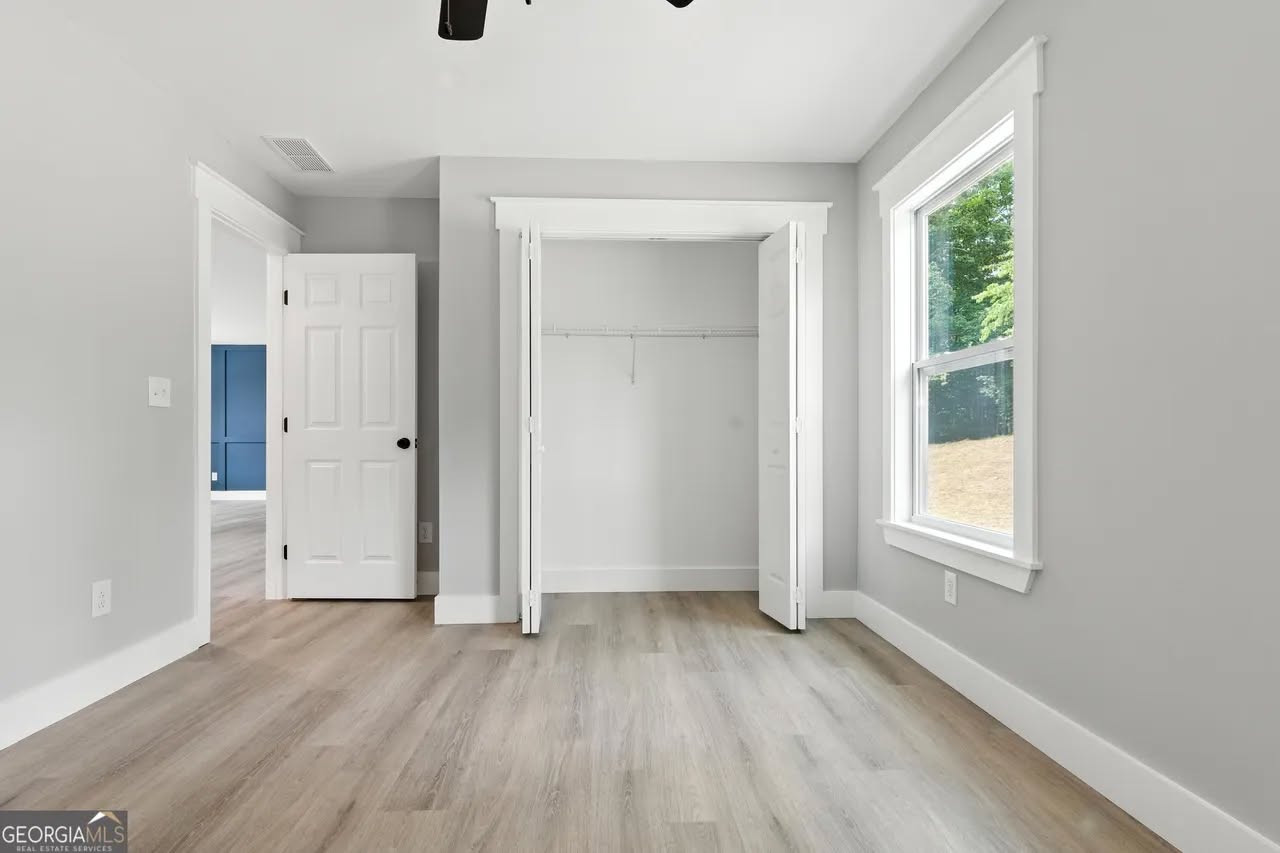 A bright, empty room with light gray walls, wood flooring, and a large window. The open closet has white double doors, creating a fresh and airy feel.