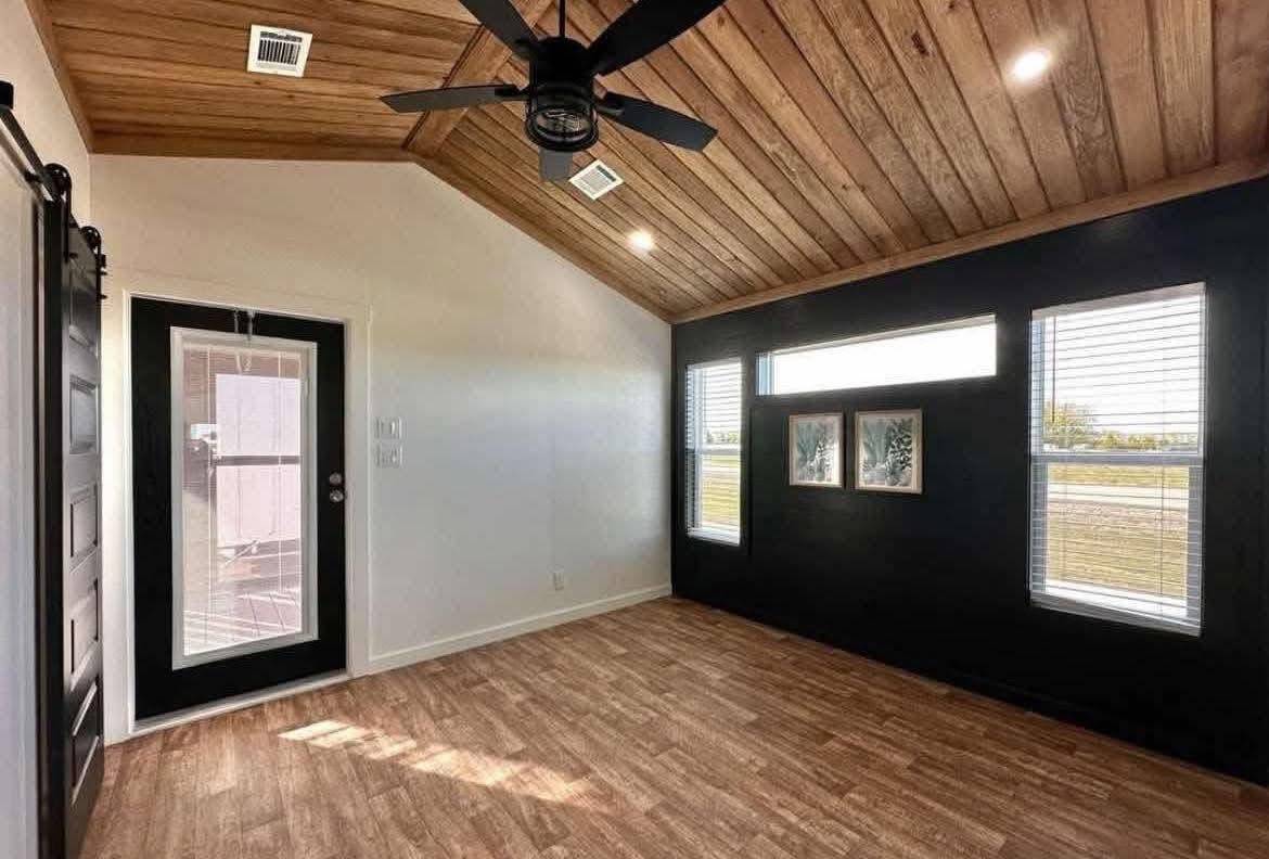 Interior of a modern room with a wooden ceiling, fan, and light wood flooring. Black accent wall with three windows and framed pictures, creating a warm, inviting atmosphere.