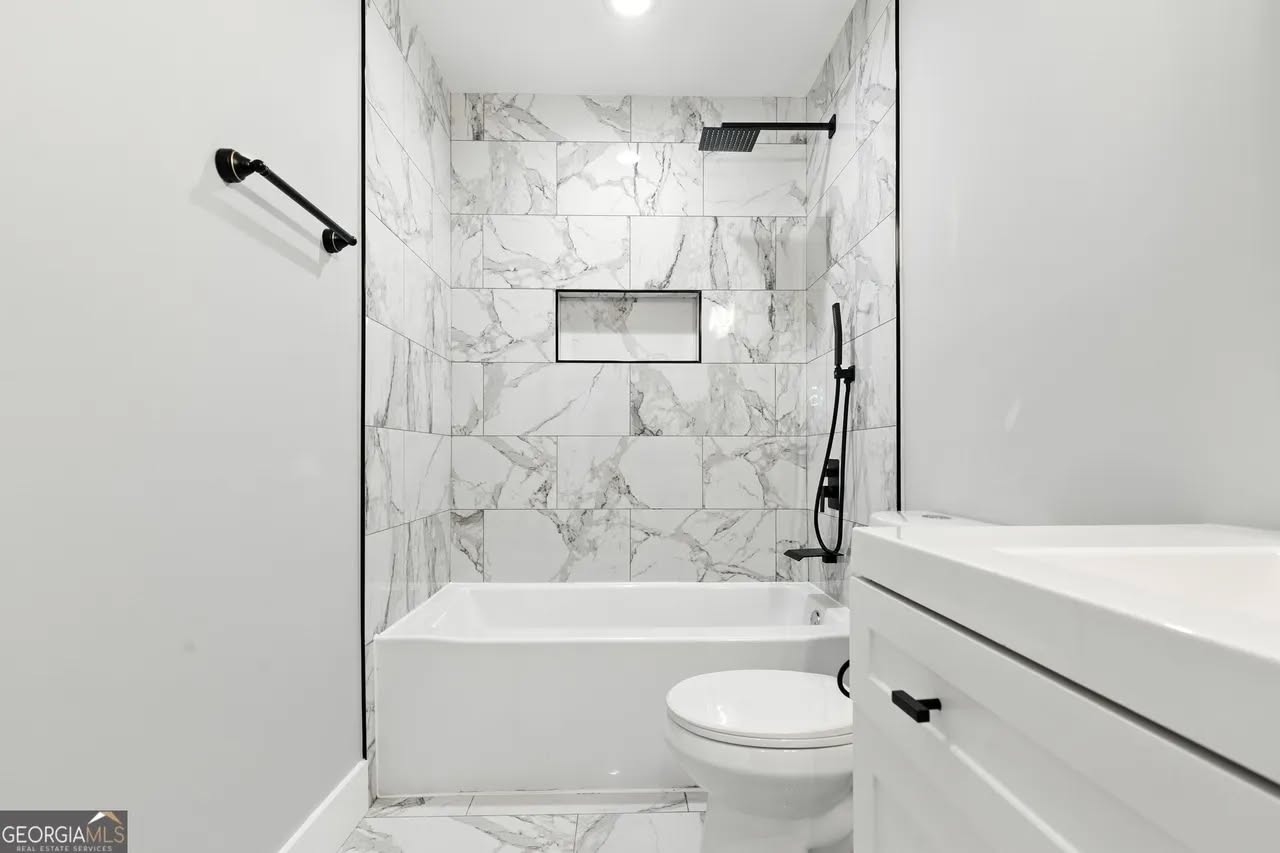 Sleek bathroom with white marble tiles, black fixtures, and a modern showerhead. Features a bathtub, toilet, and a small sink, creating a clean, minimalistic look.