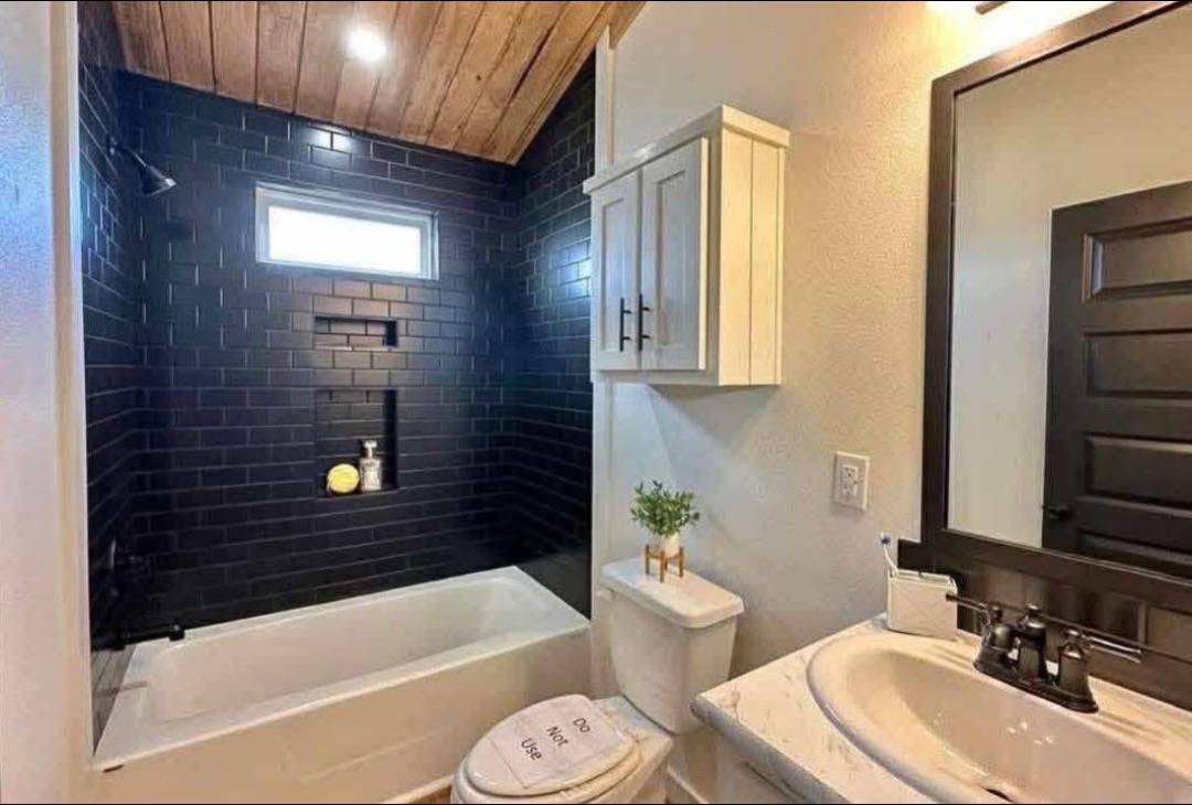 A modern bathroom features black brick tiles with a small window above a white bathtub. A white cabinet is above the toilet, which has a humorous do-not-use message. The sink is set in a light-colored countertop with a black-framed mirror, creating a clean and stylish ambiance.
