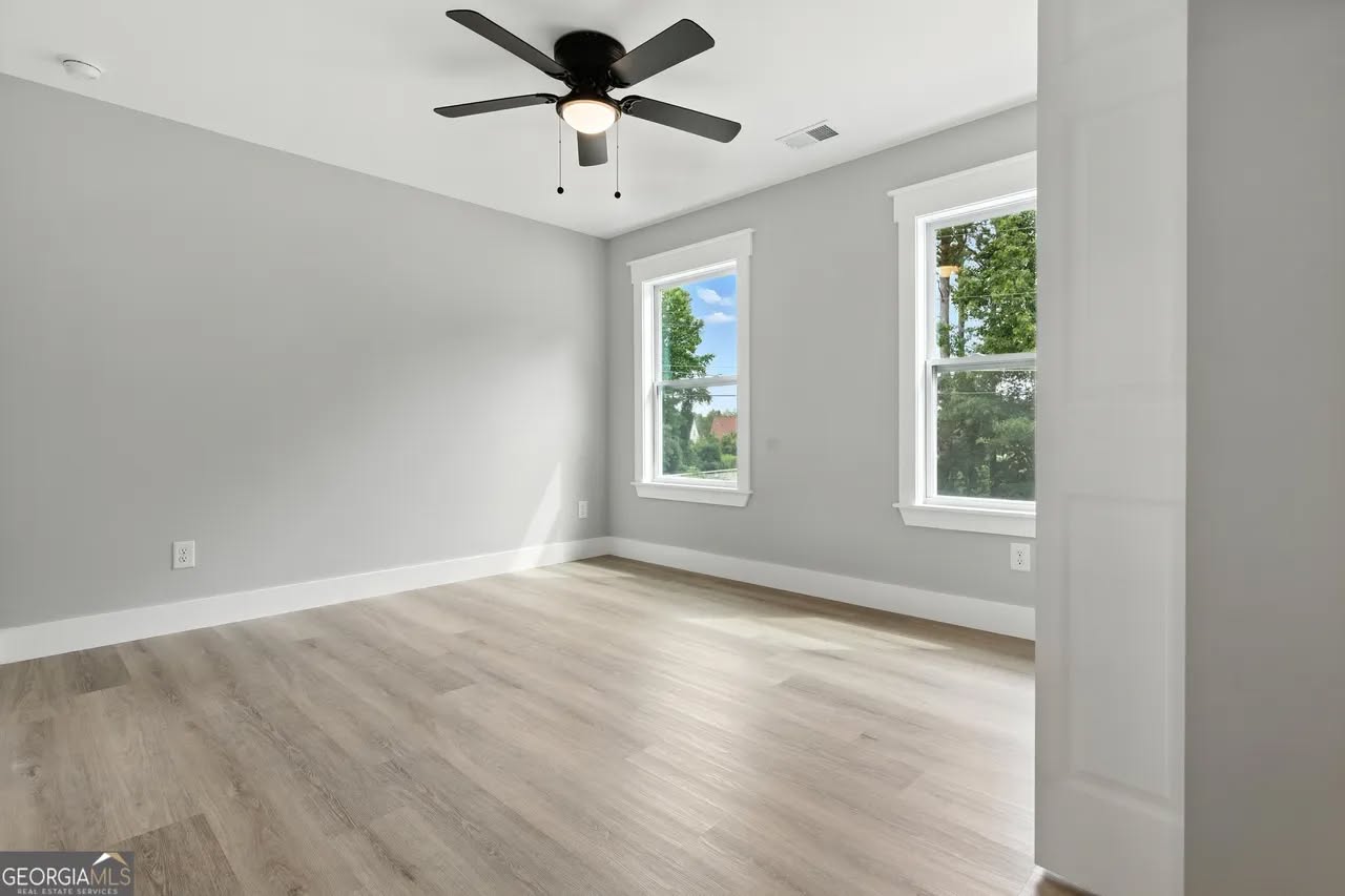 A bright, empty room with light gray walls and wood flooring. Two windows allow natural light, and a black ceiling fan adds modern elegance.