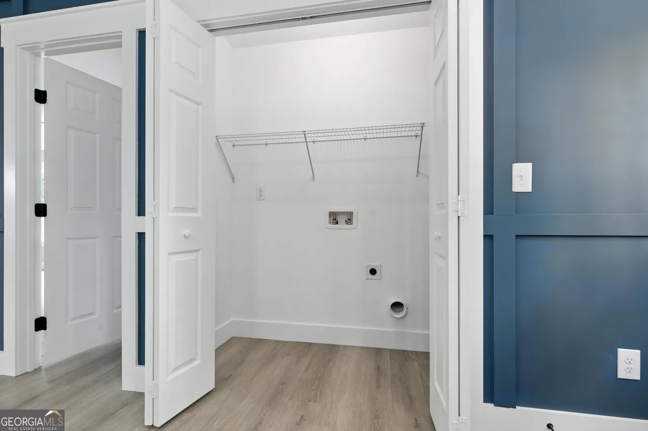 Empty laundry area with open white bifold doors, featuring a wire shelf, plumbing connections, and a smooth, light wood floor. Walls are blue and white.