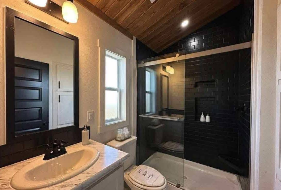 A modern bathroom with wood ceiling, black-tiled shower, glass doors, and white fixtures. A humorous toilet lid reads "Do Not Use."