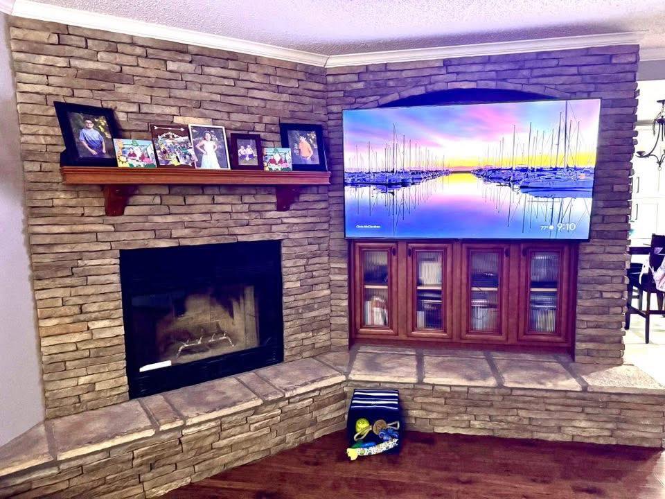 A cozy room with a stone fireplace and a mounted TV displaying a serene sunset over a marina. Family photos sit on a wooden mantle, creating a warm, inviting atmosphere.
