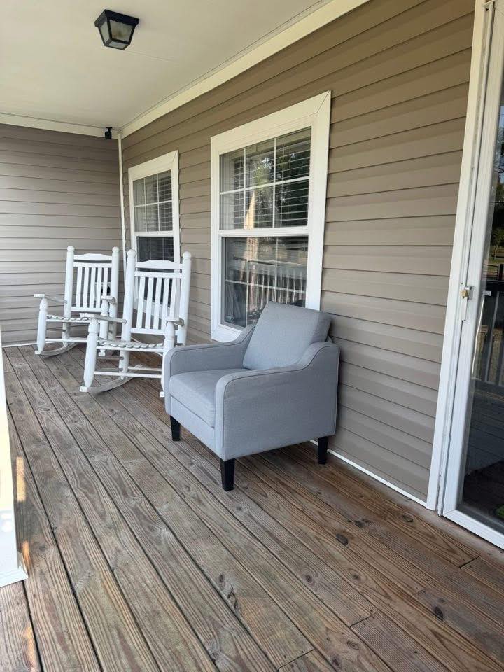 A cozy porch with wooden flooring features a soft gray armchair and two white rocking chairs. Beige siding and large windows add a calm, inviting feel.