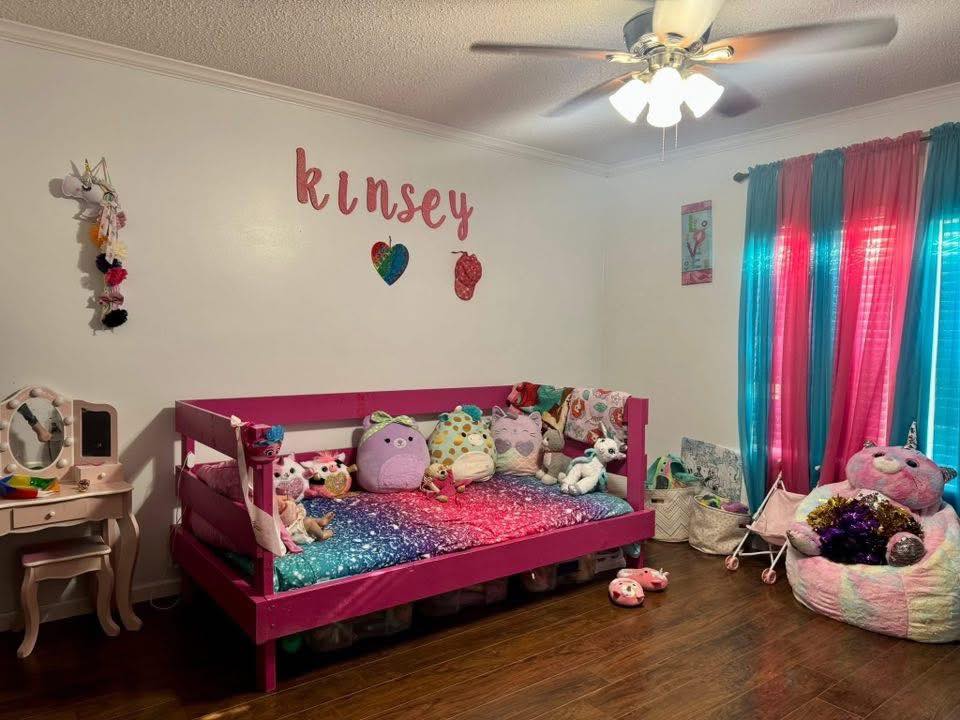 A colorful child's bedroom with a pink bed covered in stuffed animals, rainbow curtains, and a vanity. The name "Kinsey" is on the wall, creating an inviting and playful atmosphere.