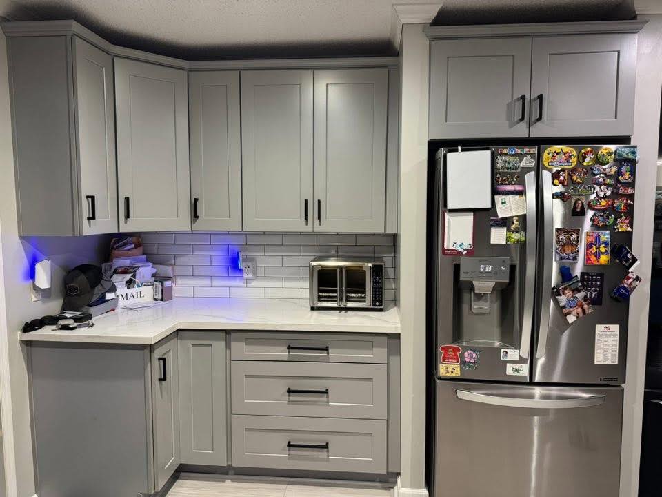 Modern kitchen with gray cabinets, stainless steel fridge with colorful magnets, and a white subway tile backsplash. A toaster oven is on the counter.