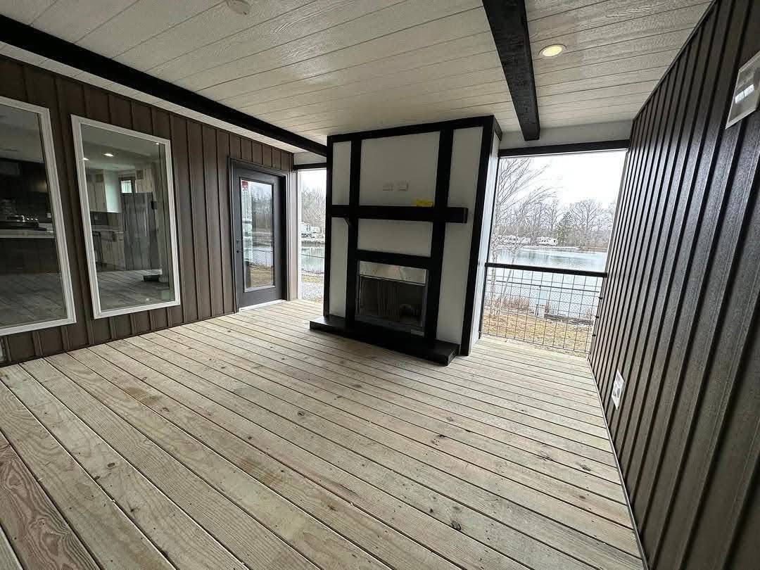 Covered patio with wooden decking and a modern black-and-white fireplace. Large windows and glass door offer a view of a serene, wooded lake area.
