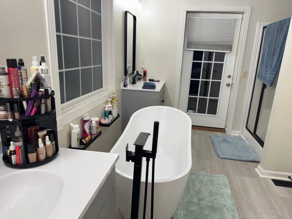 Modern bathroom with white freestanding tub, black fixtures, large vanity with mirror, and glass-paned door. Toiletries are neatly arranged. Cozy ambiance.