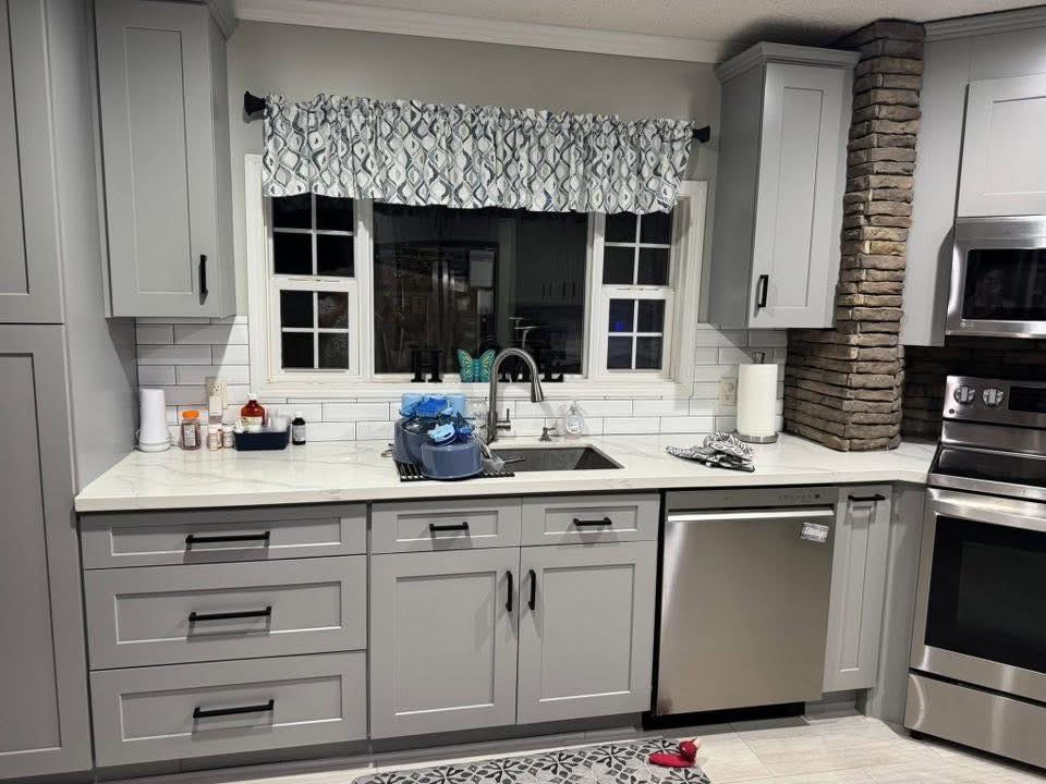 A modern kitchen with gray cabinets, a tiled backsplash, and stainless steel appliances. A window above the sink is adorned with a patterned valance.