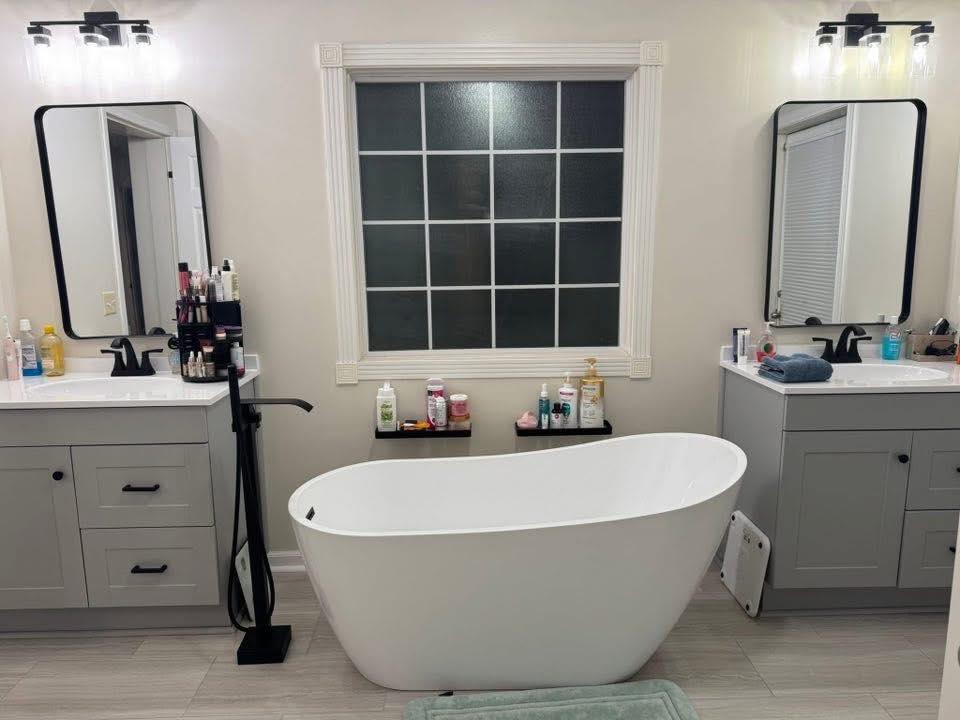 Modern bathroom with a freestanding white tub centered under a frosted window. Two vanities with mirrors, sinks, and toiletries flank the tub.