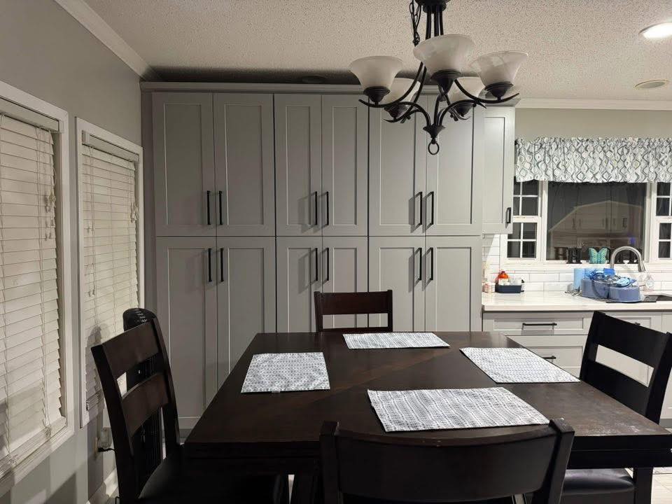 Dining area with a dark wooden table set for four, featuring light gray placemats. Gray cabinets and a window with patterned curtains are visible. A decorative light fixture hangs above. The atmosphere is cozy and organized.