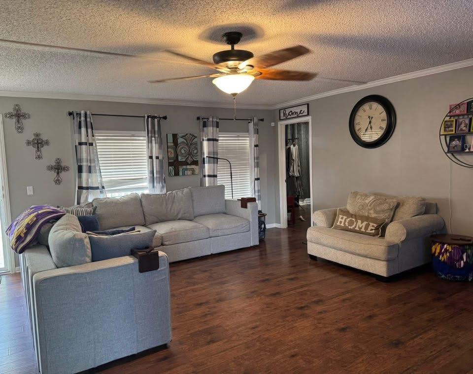 Cozy living room with gray couches, wooden floor, and a ceiling fan. Wall features cross decor and a large clock. Light, inviting atmosphere.