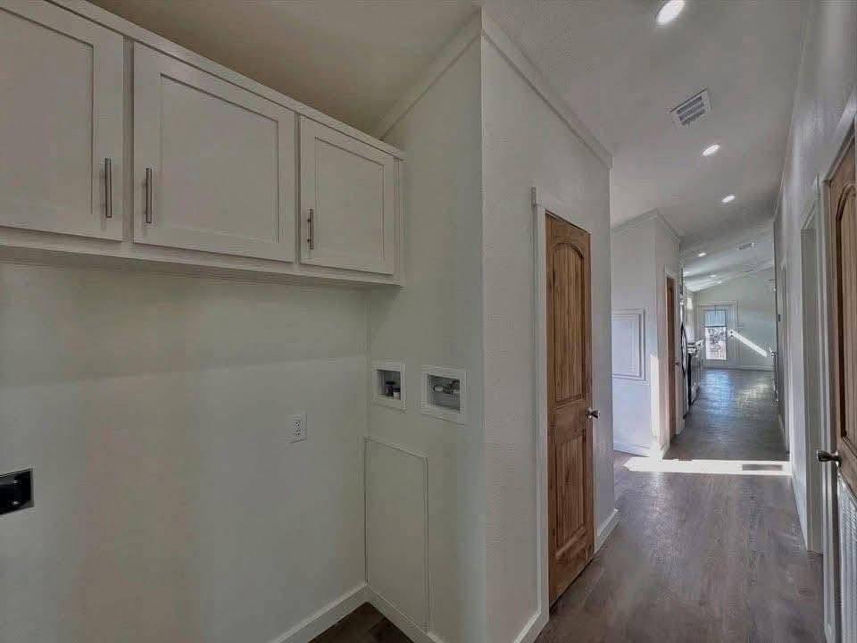 A brightly lit hallway with wooden floors and white walls. On the left, white cabinets are mounted above a utility space. Wooden doors line the right side.