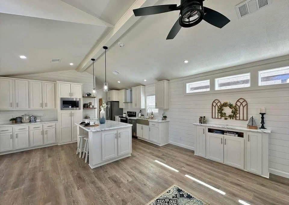 Spacious, modern kitchen with white cabinets, a central island, and wooden flooring. Pendant lights, ceiling fan, and decorative accents add warmth.