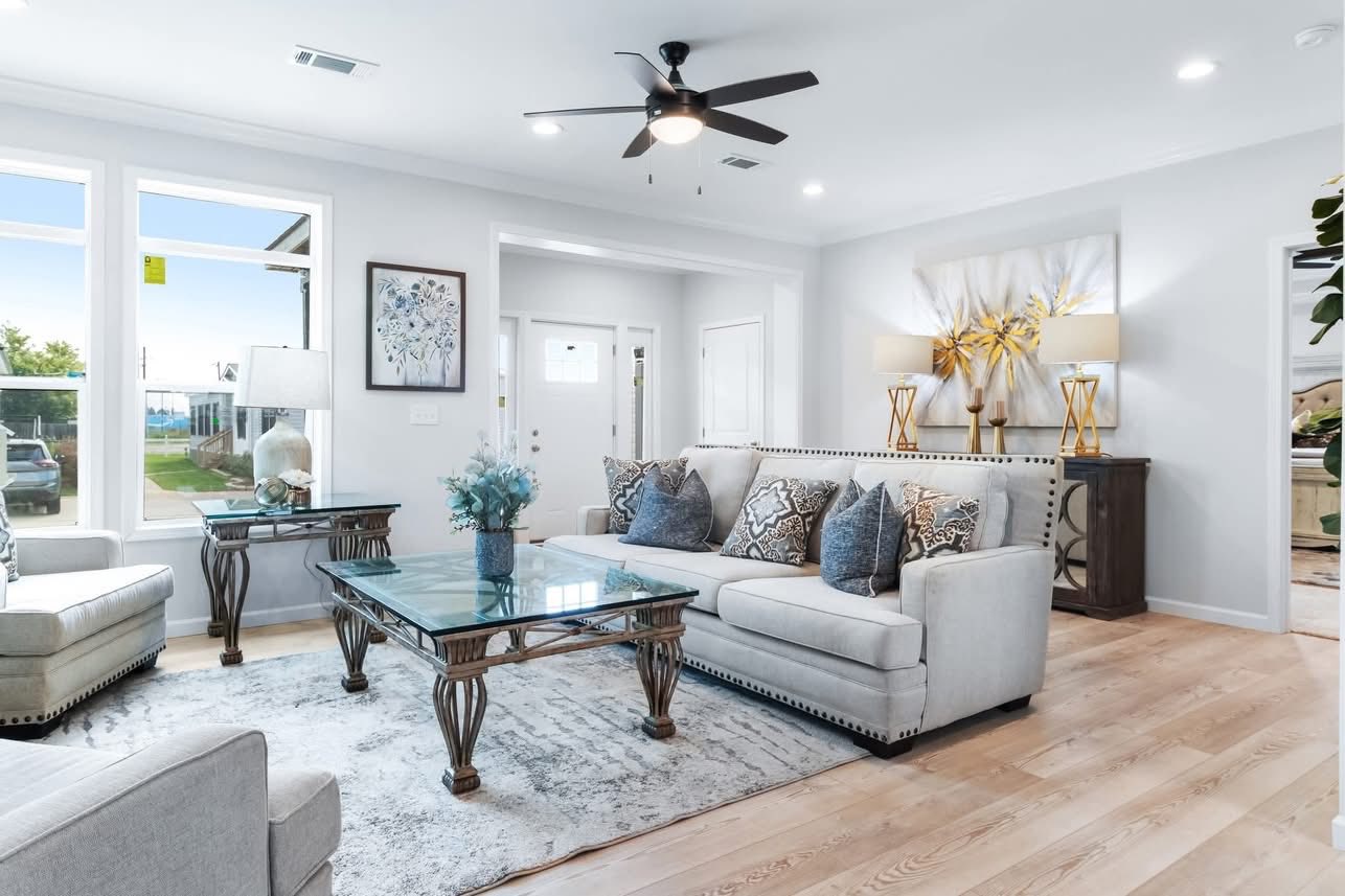 Bright, modern living room with light gray couches, a glass coffee table, and blue cushions. Art, lamps, and a ceiling fan complete the elegant look.