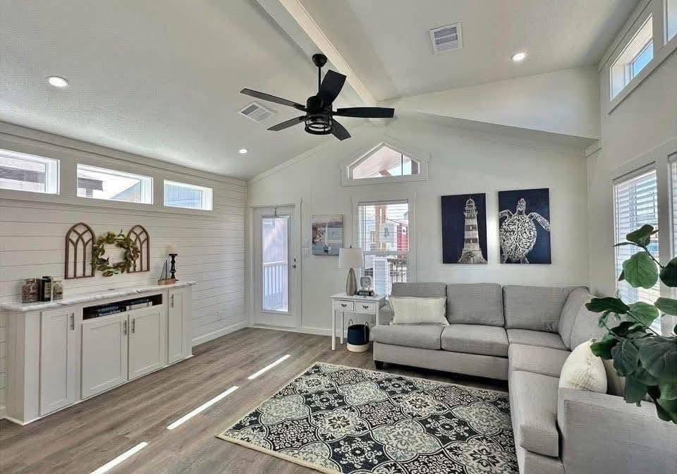 Cozy living room with a gray L-shaped sofa, patterned rug, and vaulted ceiling. Decor includes lighthouse art, plants, and a sleek ceiling fan. Bright and inviting.