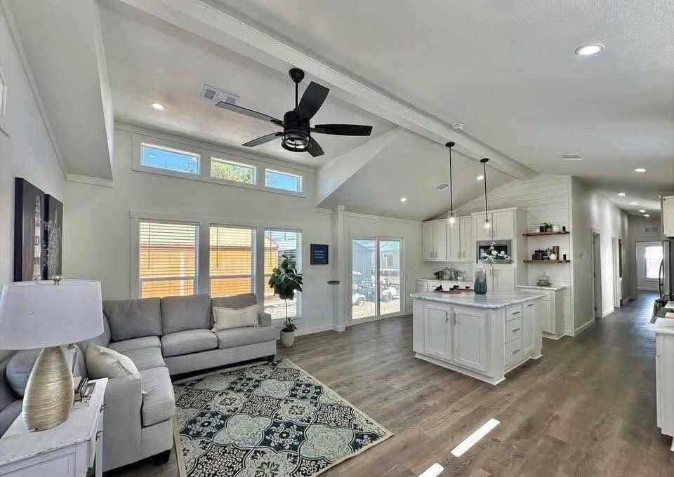 Spacious, modern living room and kitchen with high ceilings and wood floors. Features a gray sofa on a patterned rug, ceiling fan, and white cabinetry.