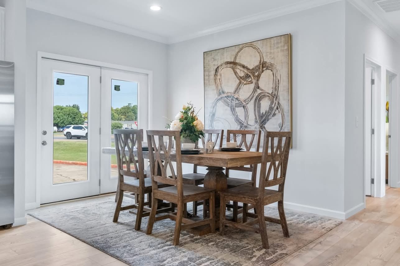 A cozy dining area with a wooden table and six chairs on a patterned rug. Large abstract painting on the wall. Glass doors reveal a green lawn outside.