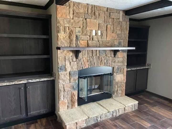 Stone fireplace with a wooden mantel is centered between two dark wooden cabinets. Reflective glass doors cover the hearth. The flooring is dark wood.