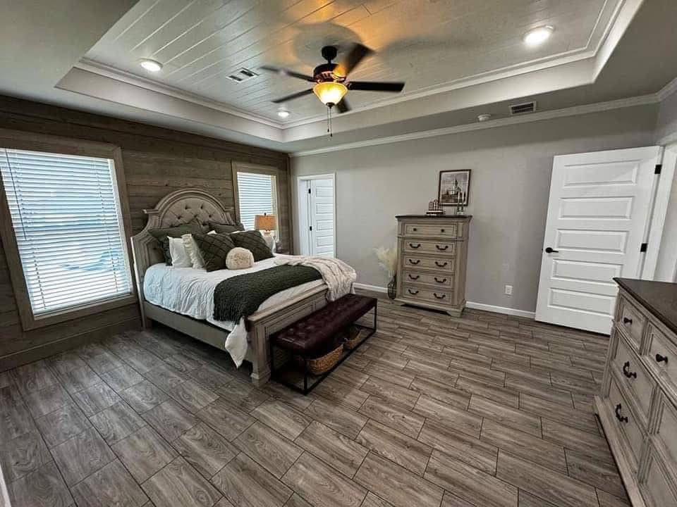 A cozy bedroom featuring a wooden bed with white and green bedding, two large windows, a ceiling fan, wooden floors, and matching dressers, creating a warm, inviting atmosphere.
