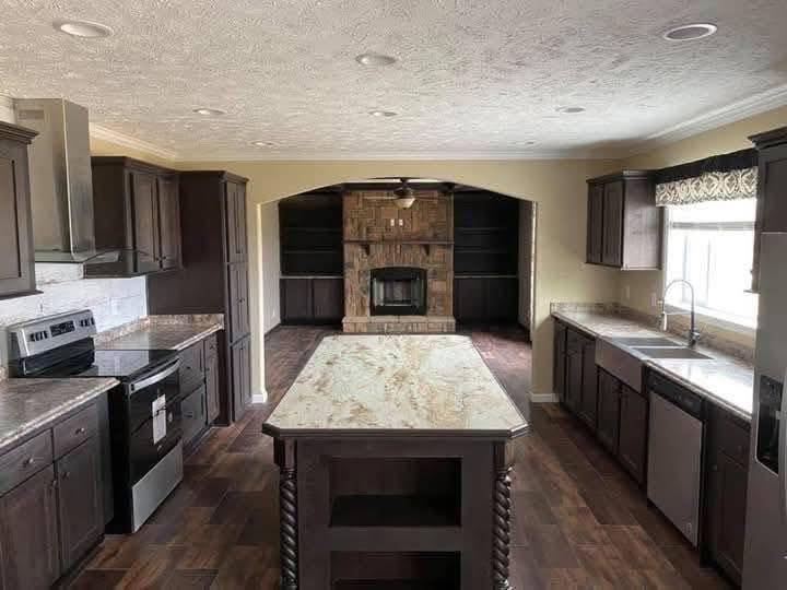 Spacious kitchen with dark wood cabinets, marble countertops, and a central island. Large window provides natural light. Cozy stone fireplace beyond.