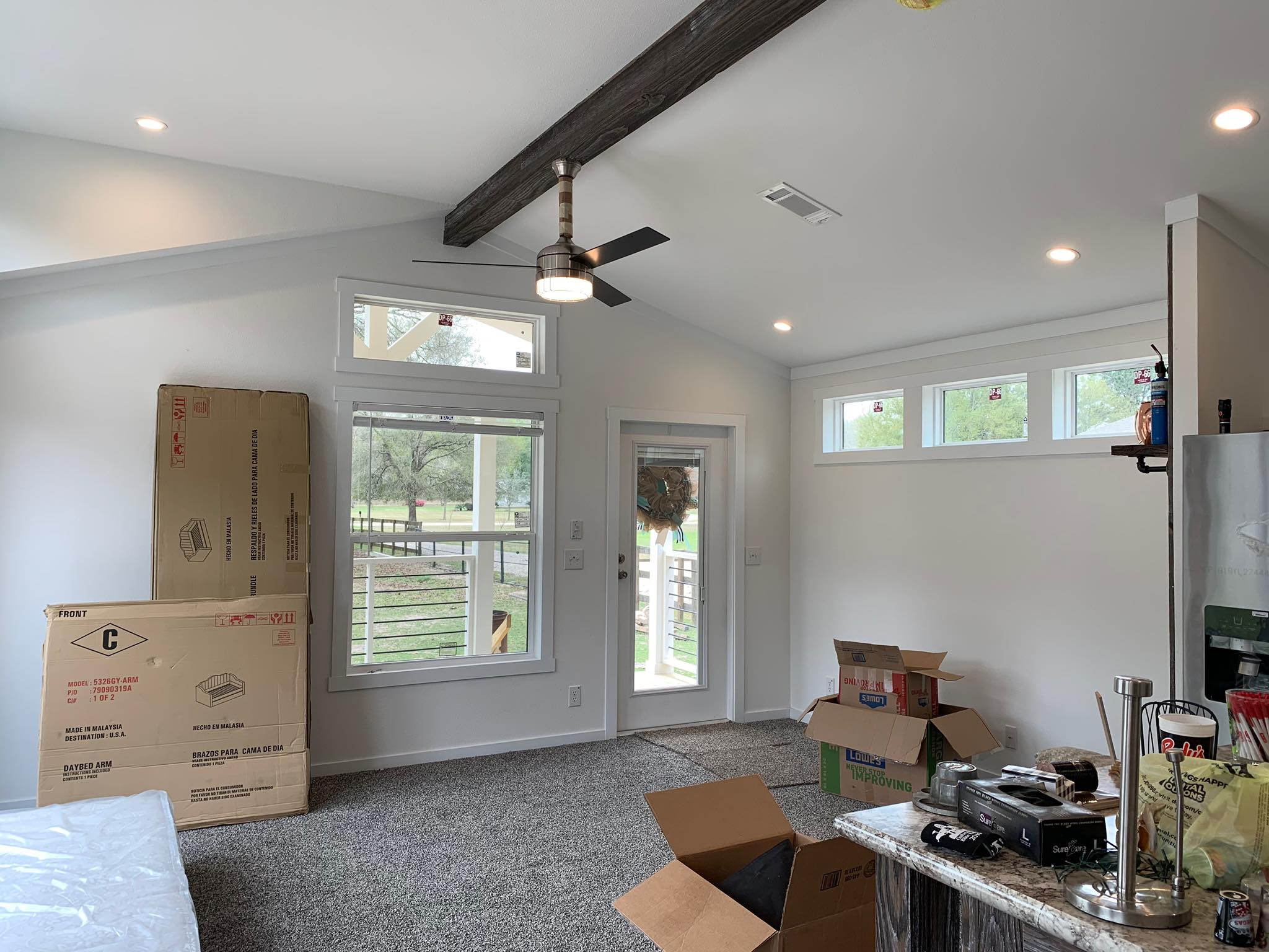 Bright, modern living room with unpacked boxes, carpeted floor, and recessed lighting. A ceiling fan adds a cozy touch, while large windows reveal a green view.