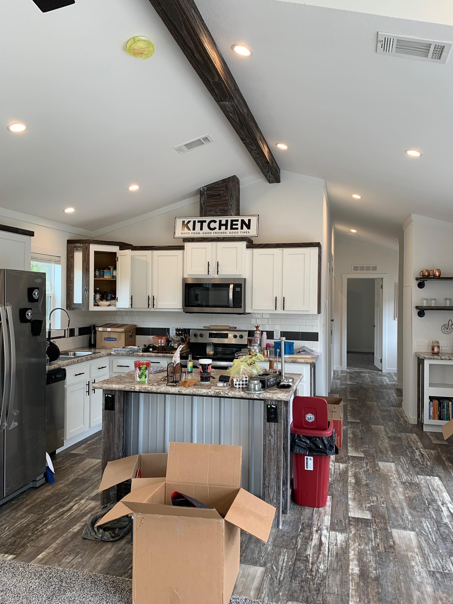 Modern kitchen with white cabinets, stainless steel appliances, and a wooden beam ceiling. A cluttered island and open cardboard box add a moving-in feel.