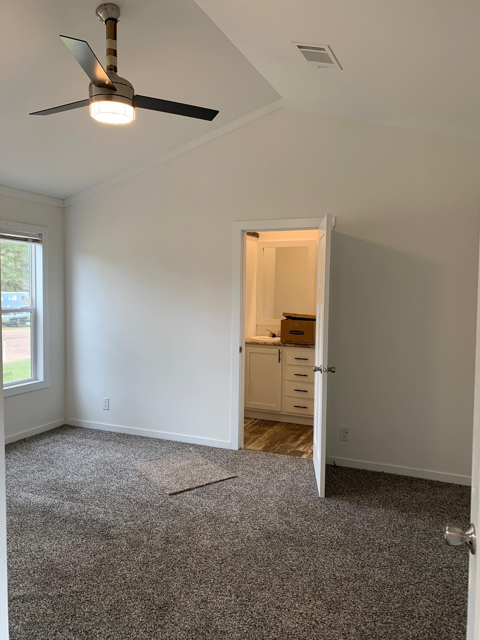A minimalist, empty room with gray carpet and white walls. A ceiling fan hangs above, and an open door reveals a bathroom with beige cabinets. Calm ambiance.