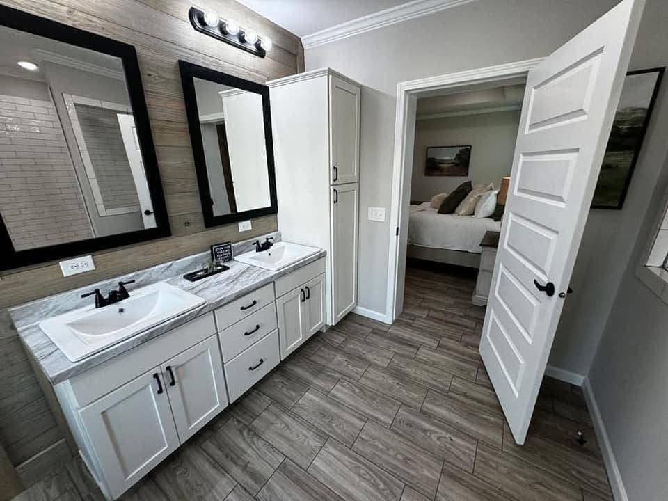 Modern bathroom with two sinks set in a white vanity, black-framed mirrors, and tiled floor. Open door reveals a glimpse of a cozy bedroom.