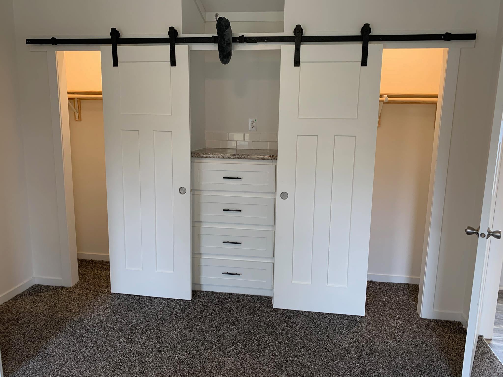 White double sliding barn doors reveal a closet with three drawers, flanked by shelving on each side. The room has a cozy carpeted floor.