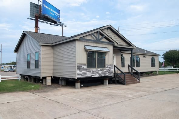 Manufactured home on concrete lot, elevated on pillars, with beige siding, brown roof, and stone accents. Nearby billboard under a cloudy sky.